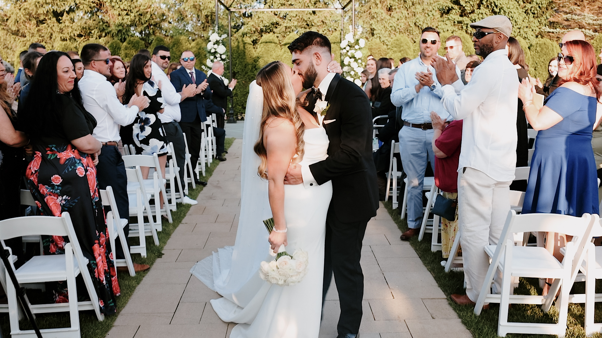 Bride and groom standing at the end of the aisle kissing. There are people standing clapping on either side of the aisle. Bride is in an elegant white dress with a long veil and her bouquet in her hand at her side. Groom is in a black tuxedo