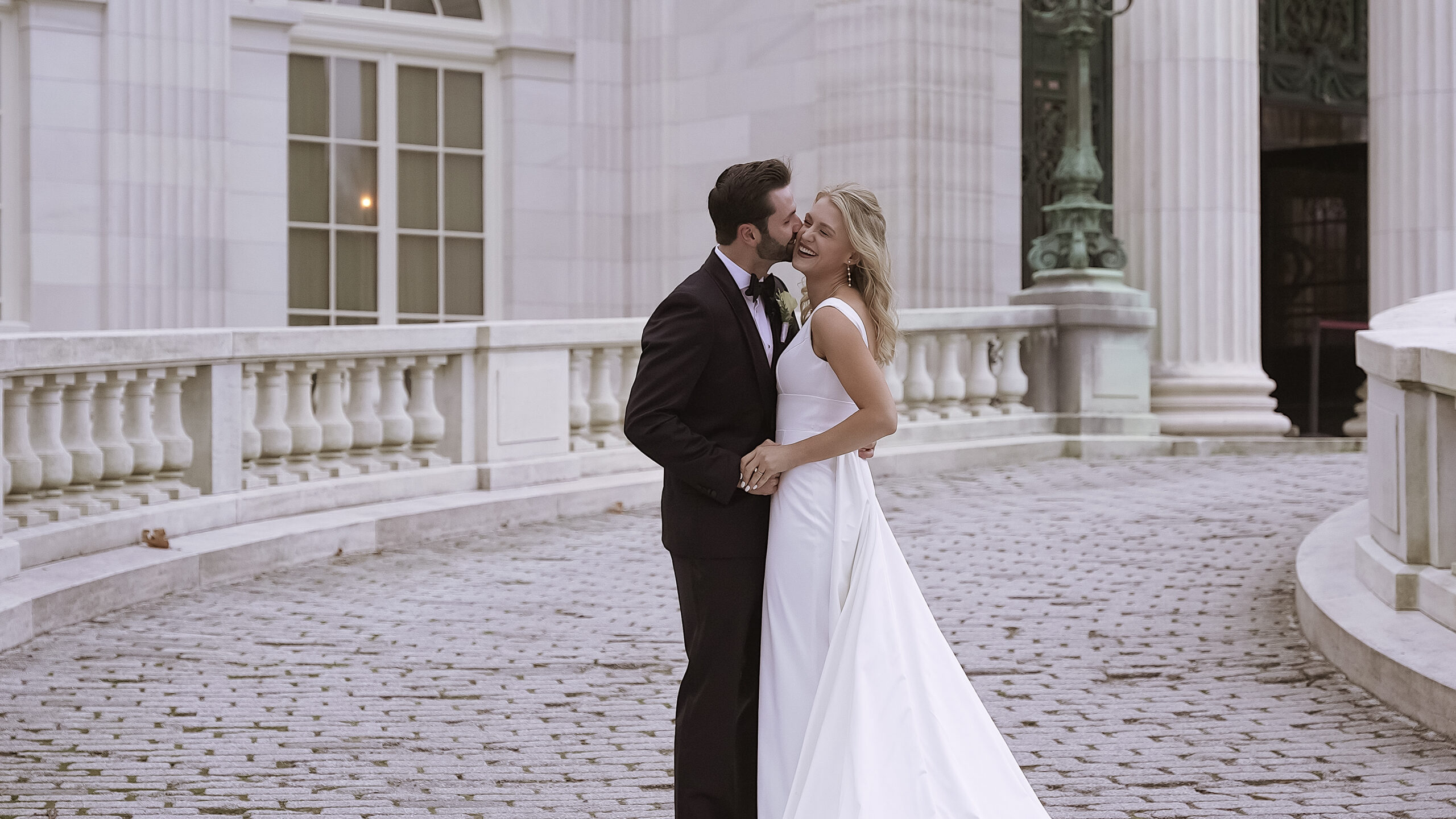 Couple standing in front of a marble building with large pillars and windows. They are facing either while standing on a cobblestone walk way. Groom is kissing bride on her right cheek and the bride is smiling. They are holding hands. Bride is wearing an elegant white dress with a removable train. Groom is wearing a black tuxedo.