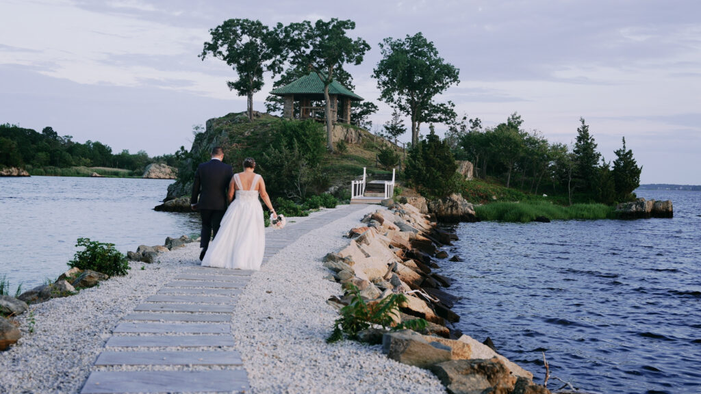 Couple walking on a stone path on a jetty toward a small island with a gazebo on top of a small hill. Bride and groom are holding hands. Bride has bouquet in her right hand and the groom is holding her left. There is water on either side of the jetty.
