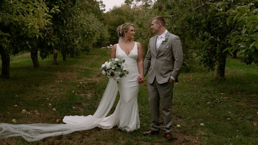 Couple standing side- by- side. holding hands in an apple orchard. Bride is wearing a low cut white dress with lace accents. She has a green and whire bouquet in her right hand and has a cathedral length veil. Groom is wearing a grey suit with a white tie and is standing with his left hand in his pocket