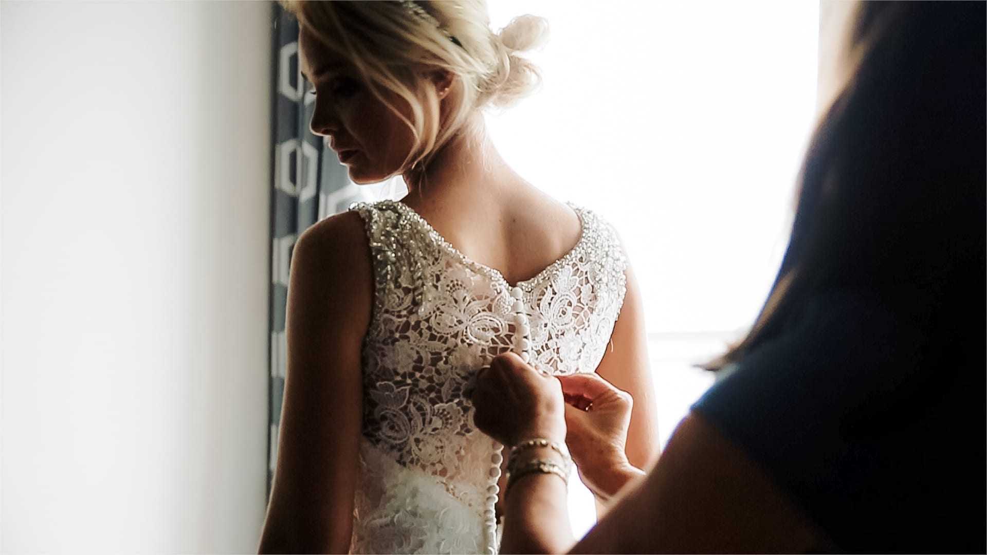 Blonde woman facing a large wedding looking over her left shoulder. Her mother is behind her finishing buttoning the back of the brides lace dress.