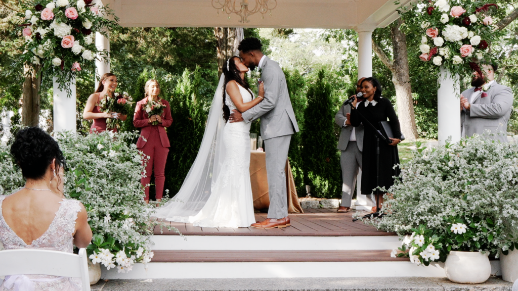 Bride and groom kissing under a gazebo. The bridesmaids are to the right in mauve. The groom and groomsmen are wearing light grey suits. The officiant is standing to the right pronoucing them. You can see a woman in a lavendar colored low back dress and dark hair in a bun clapping to the left. Bride is wearing a sleevless dress with a veil. You can see arrangements with roses on each pillar of the gazebo and on the starts are pots of green bushes and daisies.