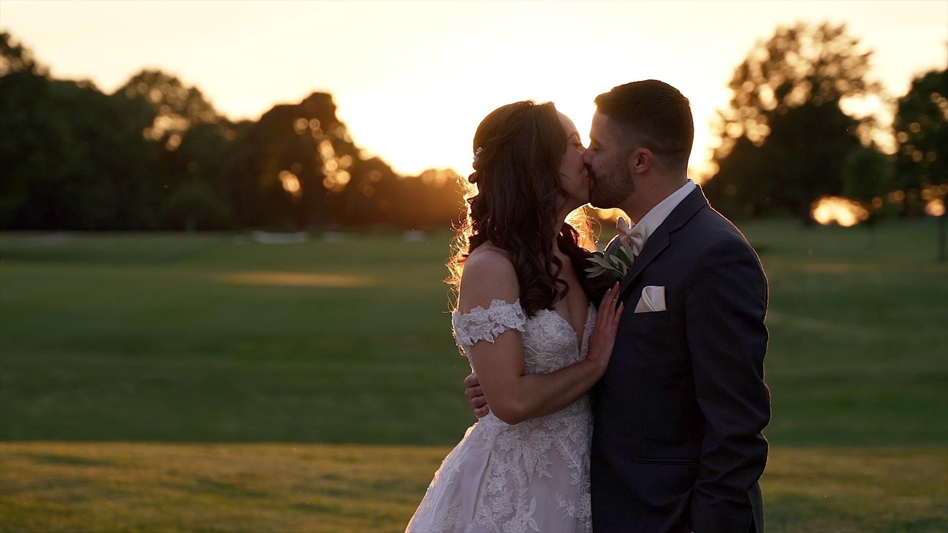 White couple kissing in front of a sunset. The sun is setting behind the trees. The groom has one arm around the bride's waist. The groom is wearing a grey suit with a pink bow tie. Bride is wearing an off the shoulder dress made of lace