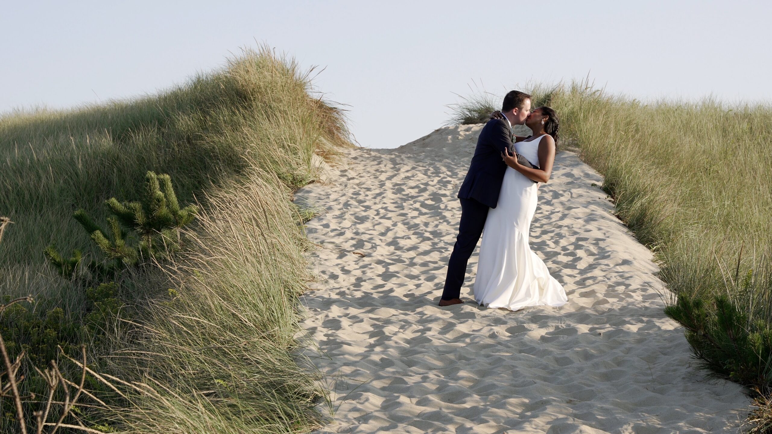Couple standing in a sandy path with American Dune Grass on either side. The groom is gently dipping the bride while kissing