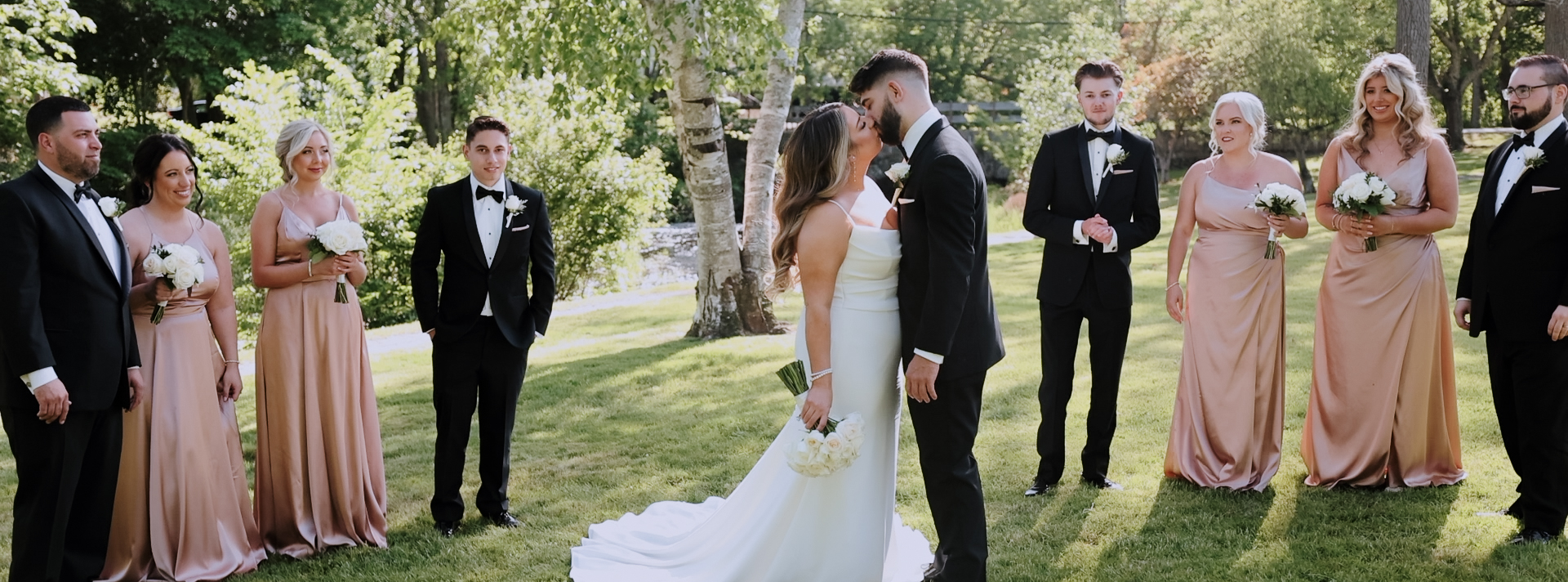 Couple kissing each other while their wedding party standing on either side of them. Bridesmaids are wearing pink satin dresses and groomsmen are wearing black tuxedos. Bride is wearing a simple but elegant wedding dress. The train of her dress is spread out over the grass. Her white bouquet with long green stems are in her right hand hanging at her side